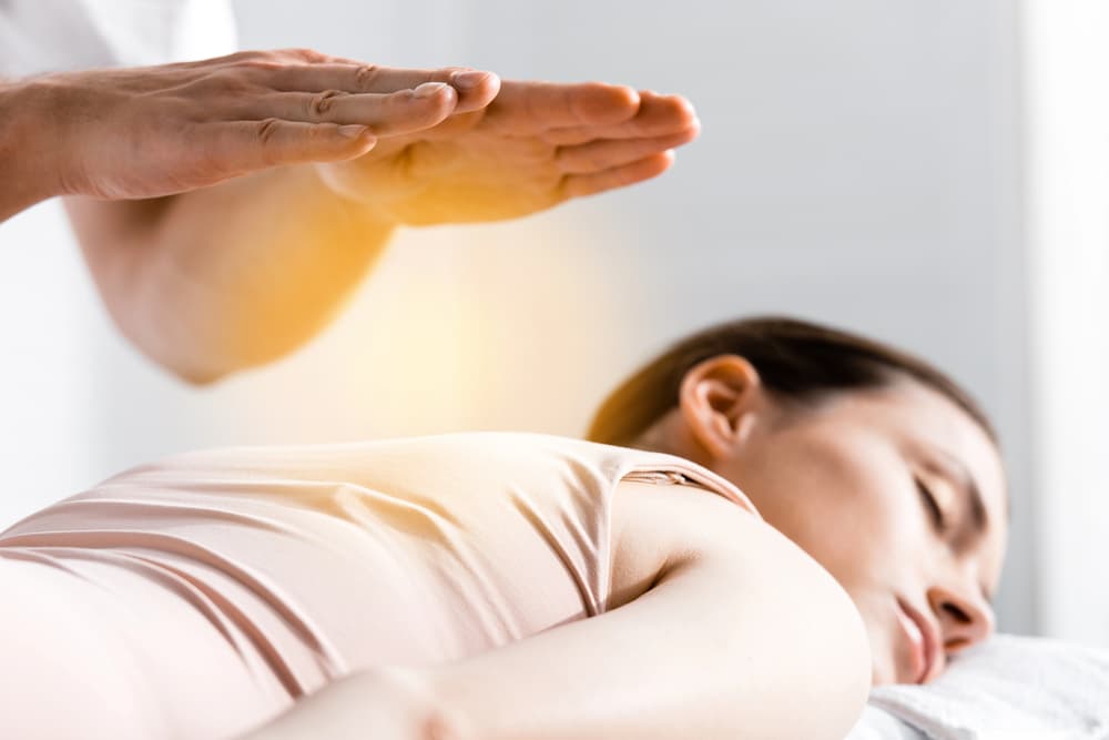 A person is lying on their stomach on a massage table, eyes closed, while another person's hands hover above their back, emitting a soft glow, suggesting energy healing. The setting is calm and serene.