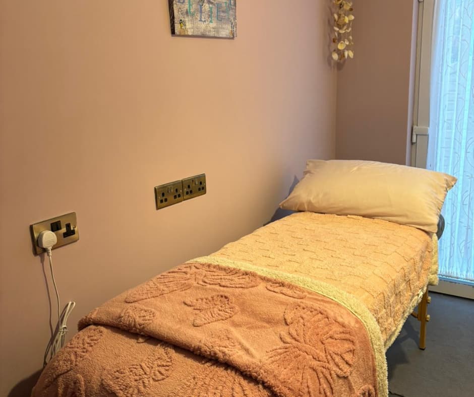 A small room with a massage table covered in a textured pink blanket and white pillow. Wall outlets and a small wall décor are visible.