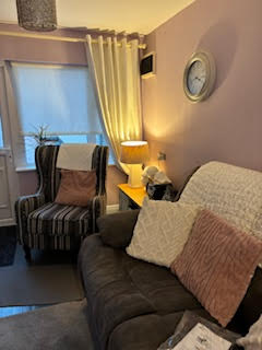 Cozy therapy room with a striped armchair and a dark sofa, both adorned with cushions. A lit lamp sits on a side table beside the sofa. A round wall clock hangs above on the lavender-painted wall. Soft curtains frame a window.